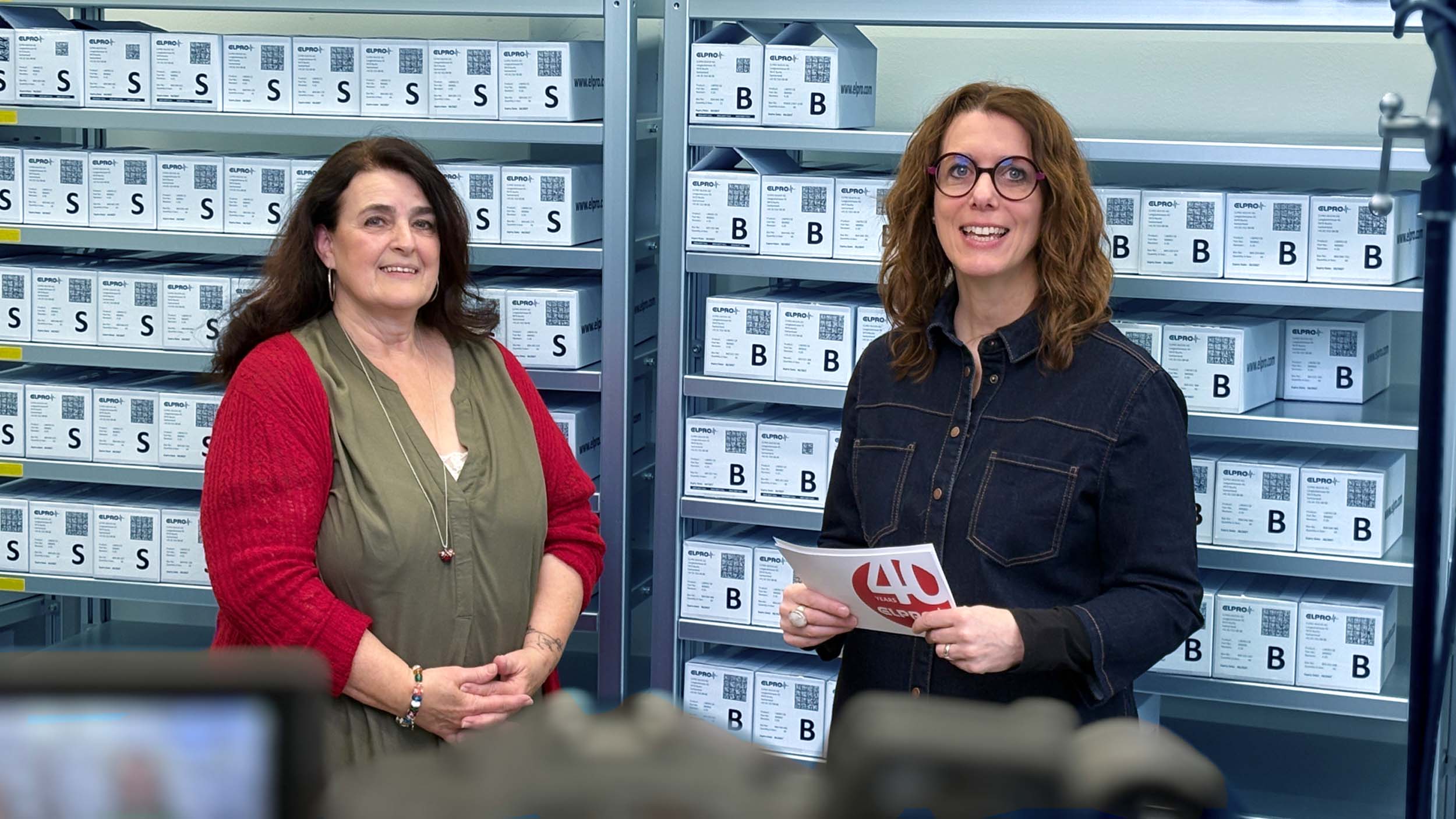 Two women are standing in front of a shelf with boxes and are looking openly and friendly at the camera.