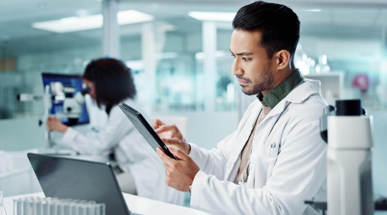 Laboratory professional using a tablet and laptop in a modern laboratory, with microscopy work taking place in the background.