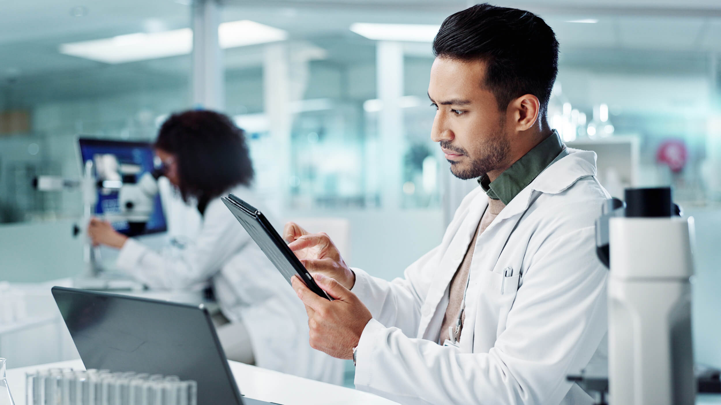 Ein Laborfachmann, der in einem modernen Labor ein Tablet und einen Laptop benutzt, während im Hintergrund mikroskopische Arbeiten stattfinden.