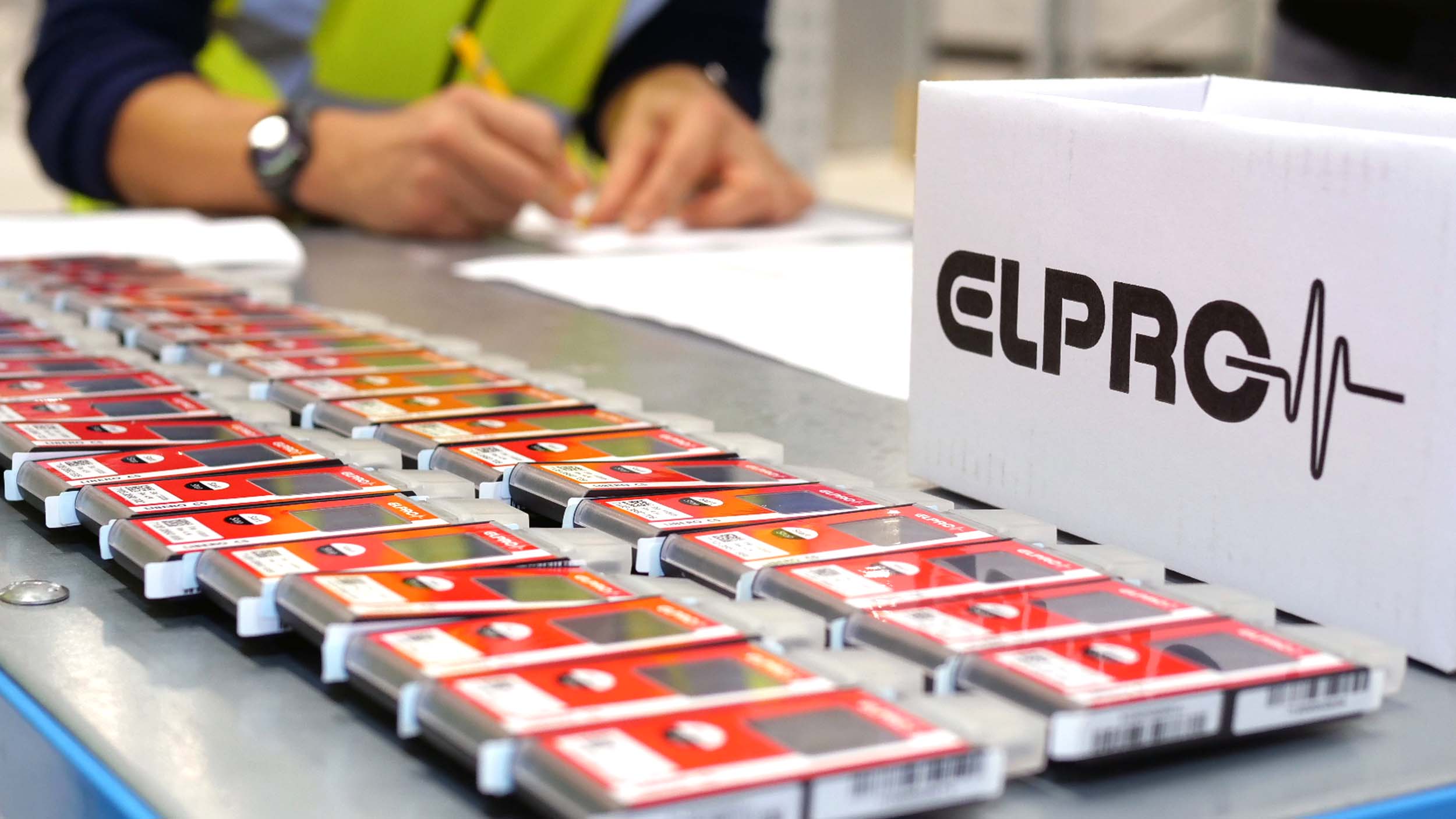 Reihen von ELPRO-Datenloggern sind auf einer Werkbank neben einer ELPRO-Box angeordnet, während eine Person im Hintergrund Unterlagen ausfüllt. Das Bild zeigt den Vorbereitungs-, Konfigurations- oder Qualitätskontrollprozess für Geräte zur Temperaturüberwachung.