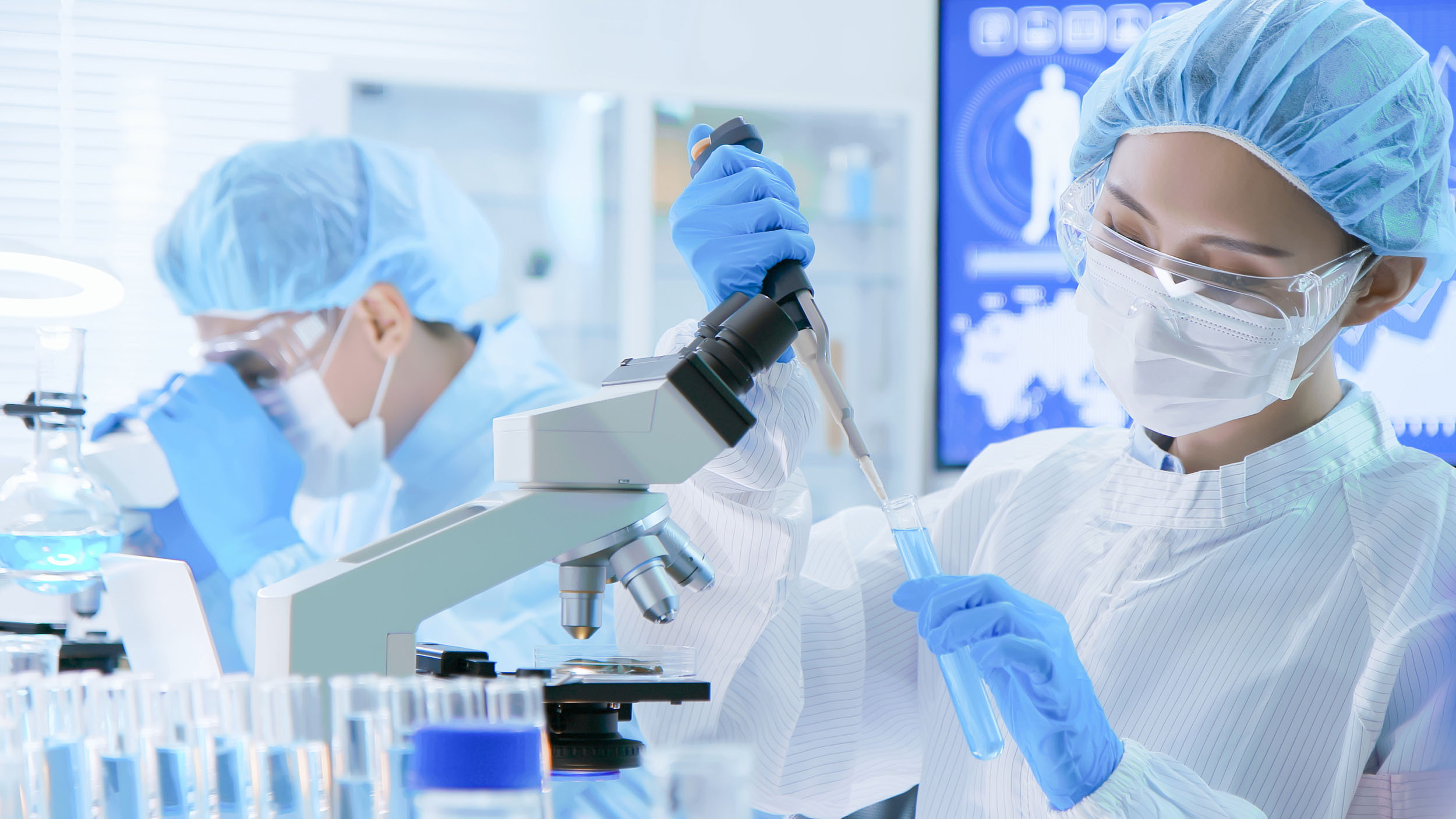 A scientist in a full white cleanroom suit and blue gloves uses a pipette in a modern pharmaceutical laboratory.