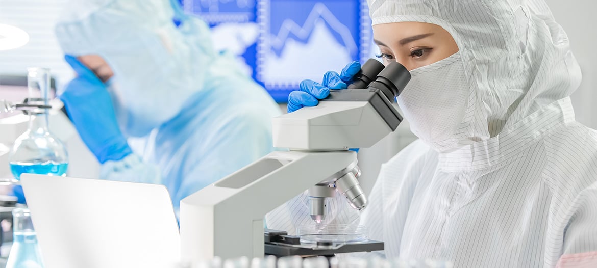 Scientist in a cleanroom suit and gloves examining a sample under a microscope in a laboratory, with another researcher and lab equipment in the background.